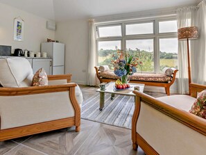 A cozy living room with a sofa set, a dining table with fruit, and a view of the outdoors through large windows. - 1 Bed in Liskeard (oc-95182) (Looe)