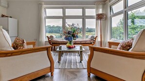 Ein Zimmer mit zwei Holzsesseln, einem kleinen Tisch mit Blumenvase und Blick auf Grün durch große Fenster.