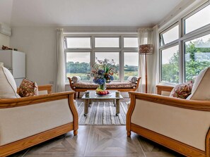 A room with two wooden armchairs, a small table with a vase of flowers, and a view of greenery through large windows. - 1 Bed in Liskeard (oc-95182) (Looe)