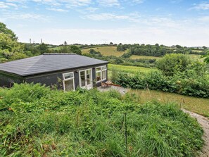 A small house with a dark roof and white-framed windows, surrounded by lush greenery and a hilly landscape.