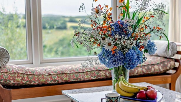 Ein Zimmer mit Glastisch, Blumen in einer Vase, Obstschale und Blick durch ein Fenster.