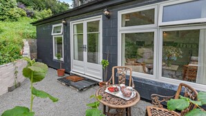 Ein Haus mit einer Terrassenfläche, komplett mit Tisch und Stühlen, einem kleinen Garten und Blick ins Innere durch große Fenster.