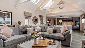 A spacious living area with a high ceiling, a large sectional sofa, a coffee table with a wine bottle and flowers, and a view of the kitchen in the background. - 1 Bed in Glasgow (oc-83392) (Eaglesham)