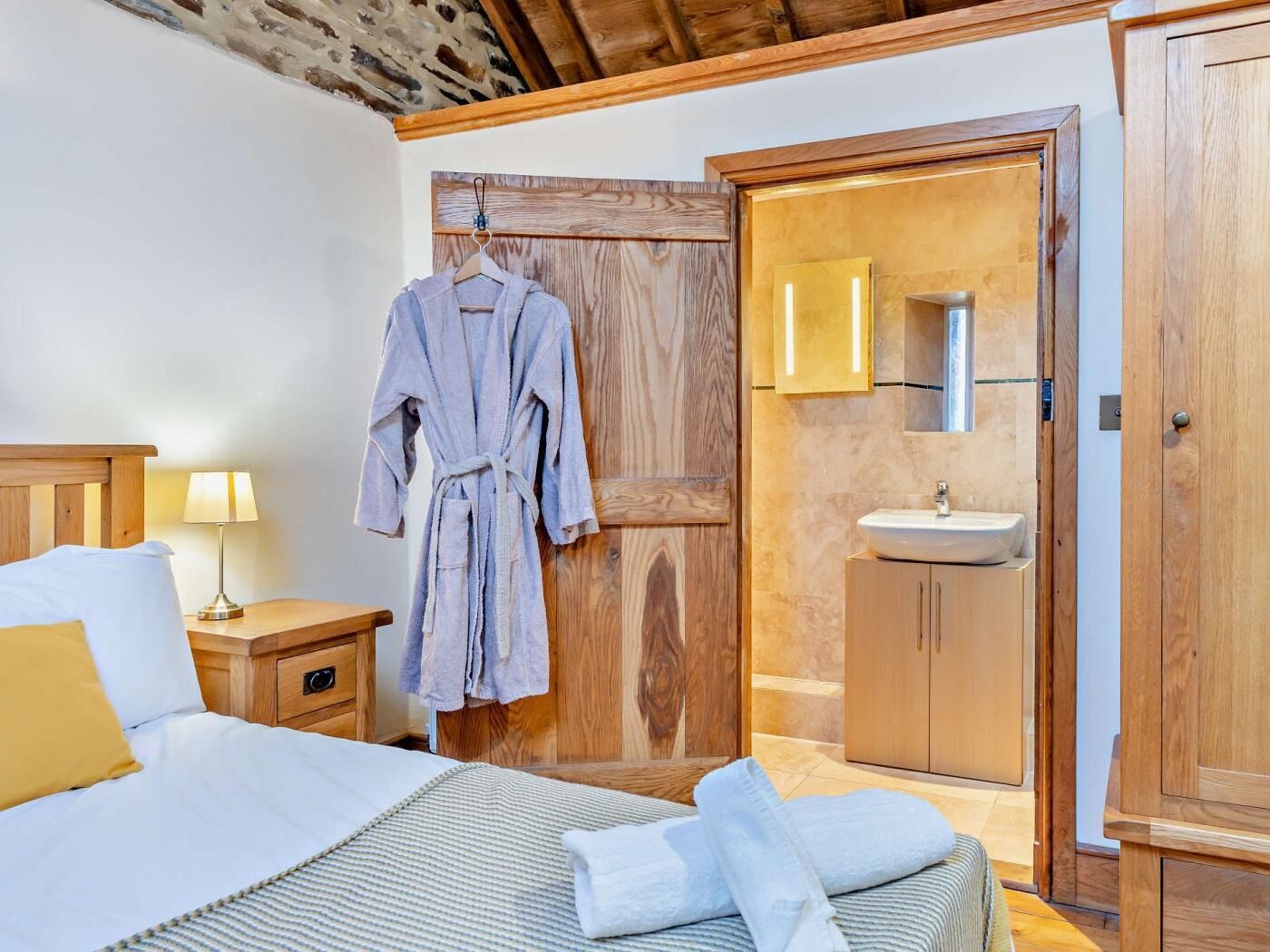 Ein Schlafzimmer mit Bett, Nachttisch, Lampe, Holztür und einem Badezimmer mit Waschbecken und Spiegel.