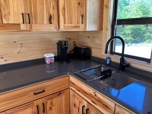 A kitchen with wooden cabinets, a black coffee maker, a kettle, and a sink with a faucet. - Cozy 1-bedroom log cabin in peaceful Gouldsboro with AC & view of Frenchman Bay (Gouldsboro)