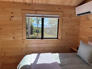 Quarto de cabana de madeira com uma cama, janela com persianas e aparelho de ar condicionado.