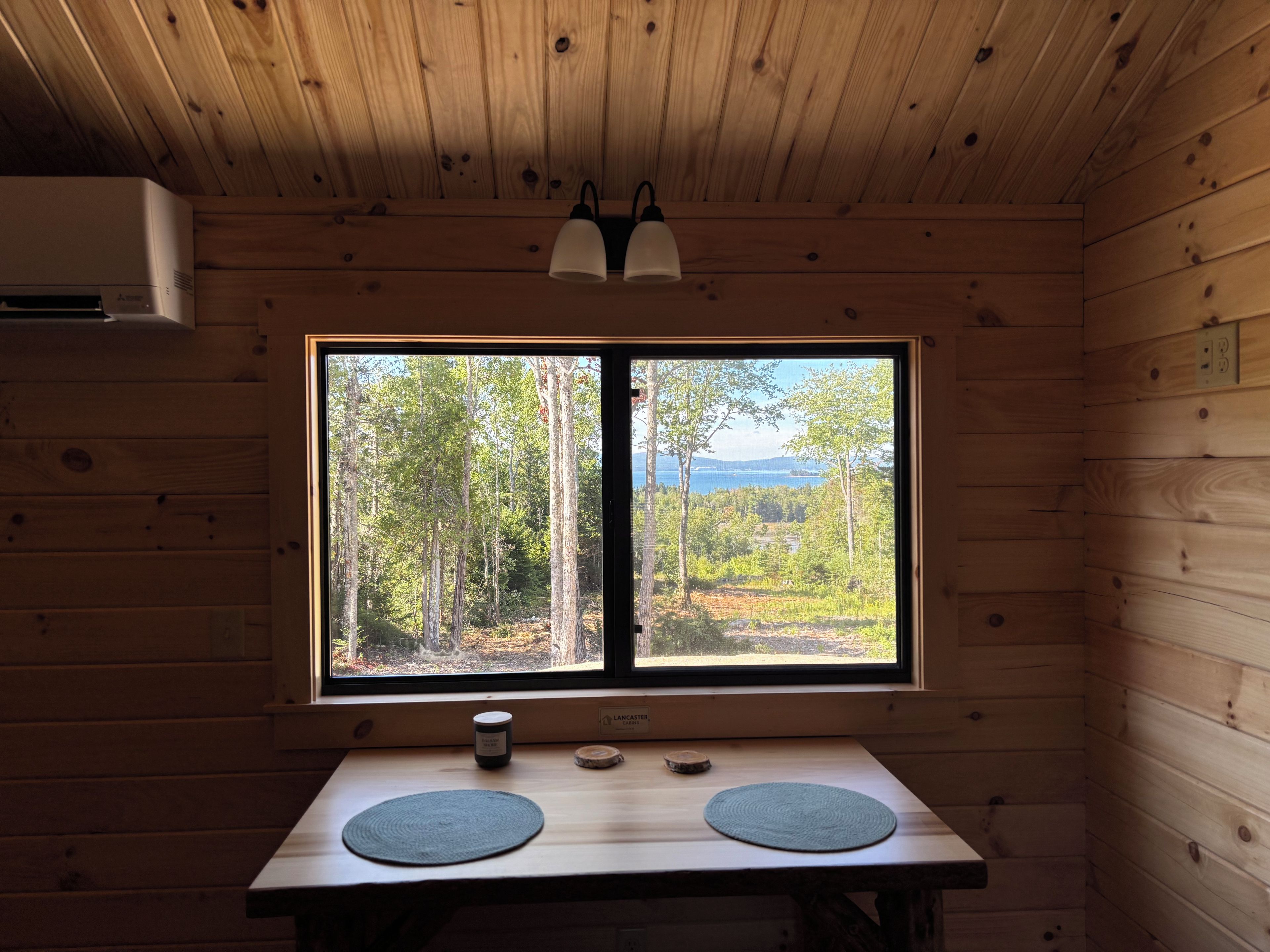 A wooden cabin interior with a large window, a table with two placemats, and a small container on the table.