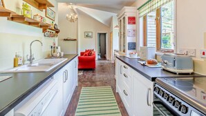 A kitchen with a stove, sink, and a red sofa in the background. - 3 Bed in Watchet (oc-95161) (Washford)
