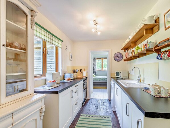 A compact kitchen with a built-in oven, a microwave, and a sink. There is a window with blinds, a clock on the wall, and a shelf with kitchenware.