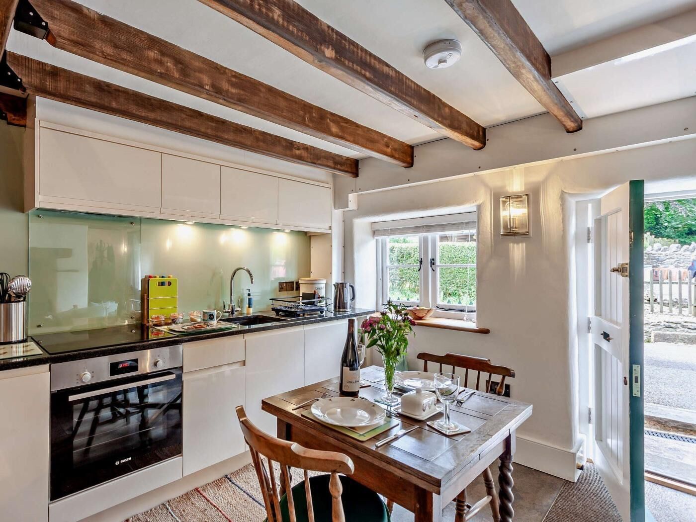 A kitchen with a Bosch oven, a dining table set for four, wooden beams on the ceiling, and a window with a view of greenery.