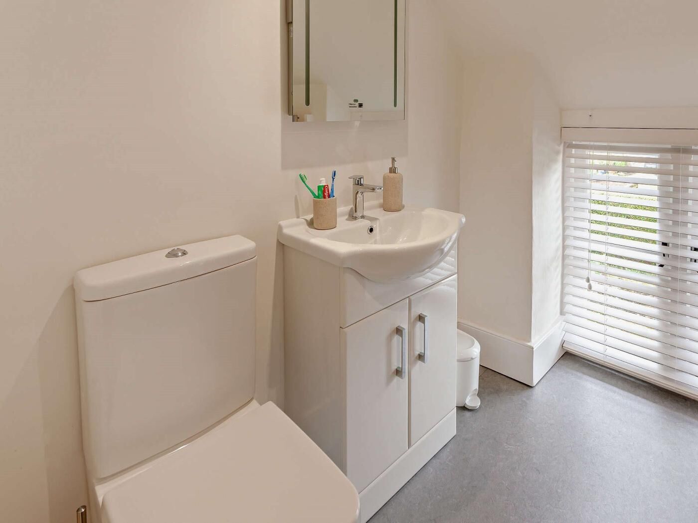 A bathroom with a toilet, sink, mirror, and window with blinds.