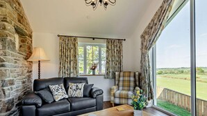 A living room with a stone wall, a black leather sofa, a wooden coffee table, and a view of a green landscape through large windows. - 2 Bed in Llanarth (oc-95322) (Llanarth)