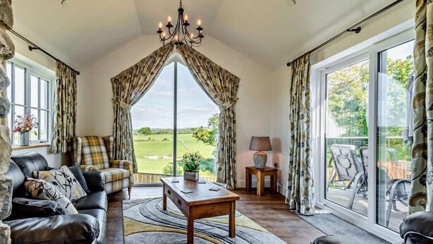 A spacious room with a large window, a chandelier, a sofa, a coffee table, and a view of the outdoors. - 2 Bed in Llanarth (oc-95322) (Llanarth)