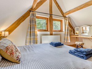 Ein Schlafzimmer mit schrägen Wänden, einem Bett mit gestreifter Bettwäsche, einem Fenster mit Vorhängen, einem Heizkörper und einem Nachttisch mit Lampe.