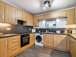 A modern kitchen with wooden cabinets, a built-in oven, a washing machine, and a countertop with a kettle and coffee maker. - 2 Bed in Holmfirth (oc-95230) (Holmfirth)