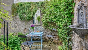 A rustic outdoor seating area with a table and chairs, a decorative fountain, and a wall with a window and ivy. - 3 Bed in Kelso (oc-95231) (Kelso)