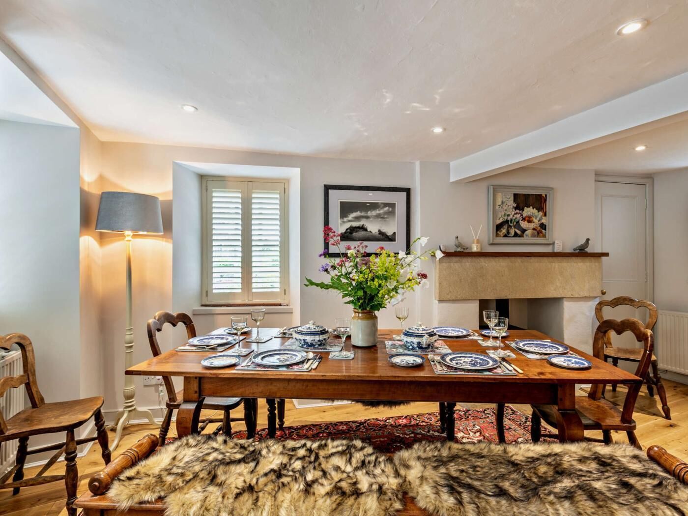 A dining room with a wooden table set for four, flanked by chairs, a fur rug, and a vase with flowers.