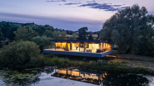 Uma casa moderna com uma extensão de vidro sobre a água, rodeada por árvores.