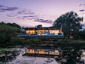 A modern house with a glass facade, situated by a body of water with reflections.