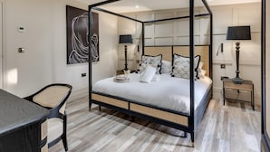 Uma cama de dossel com colchão branco, duas mesas de cabeceira com abajures, uma cadeira de vime e um quadro abstrato grande na parede.