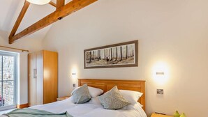 Un dormitorio con cabecera de madera, cama con ropa blanca, un cuadro de un bosque, lámparas de noche y una ventana con vistas.
