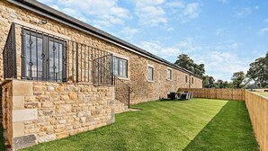 Un edificio de piedra con un patio cubierto de césped, una valla de madera y una mesa con sillas.