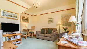 A cozy living room with a flat-screen TV, wooden furniture, a sofa with cushions, a bookshelf, and a map on the wall. - 1 Bed in Keswick (oc-87483) (Applethwaite)