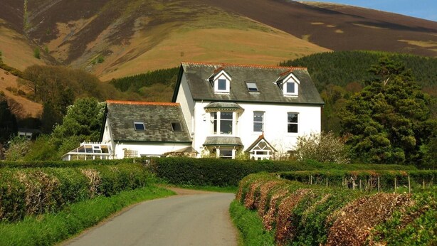 A white building with a sloped roof, surrounded by greenery and a mountain in the background. - 1 Bed in Keswick (oc-87483) (Applethwaite)