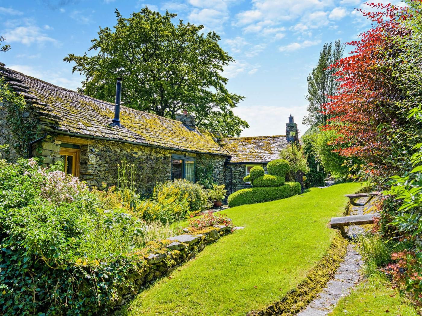 A stone cottage with a thatched roof, surrounded by a well-maintained garden with a variety of plants and a neatly trimmed lawn.