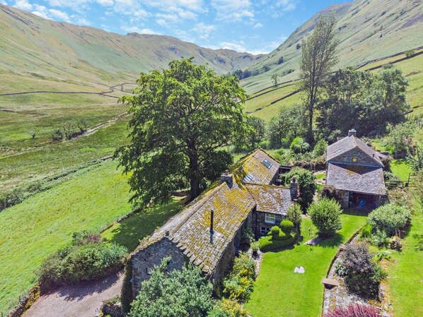 A stone house with a sloped roof, surrounded by lush green fields and a large tree. - 6 Bed in Penrith (oc-95344) (Ullswater)