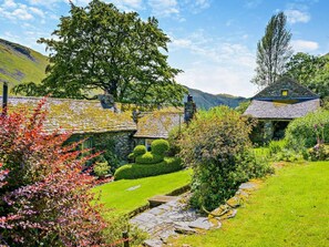 A stone cottage with a thatched roof, surrounded by a well-maintained garden with a hedge maze and a small stream. - 6 Bed in Penrith (oc-95344) (Ullswater)