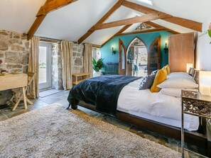 A bedroom with a stone wall, wooden beams, a bed with white and yellow pillows, a desk, and a plant. - 1 Bed in Penzance (oc-92234) (Lamorna)