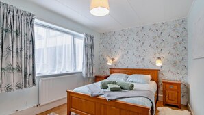 Ett sovrum med en säng i trä, en mjuk matta, en fäll, ett fönster med gardiner och en tapet med blommönster.