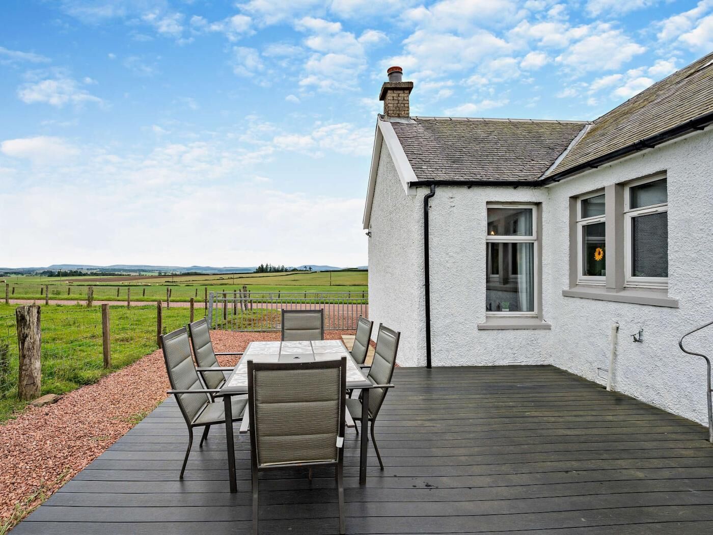 A white building with a tiled roof, a patio with a table and chairs, and a view of a green field.