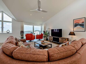 A spacious living room with a large sofa, red chairs, a TV, and a ceiling fan. - 4 Bed in Bideford (oc-95384) (Westward Ho!)