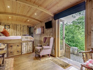 A rustic cabin interior with a wooden ceiling, a bed, a plaid armchair, and a view of greenery outside. - 1 Bed in Kendal (oc-95349) (Ings)