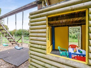 A log cabin with a yellow interior, a table with chairs, and a swing set outside. - 5 Bed in Torrington (oc-knawo) (Frithelstock)
