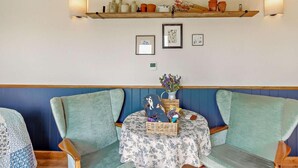 Una habitación con una zona de estar con banca de madera, una pequeña mesa con mantel floral y estantes en la pared con objetos decorativos.