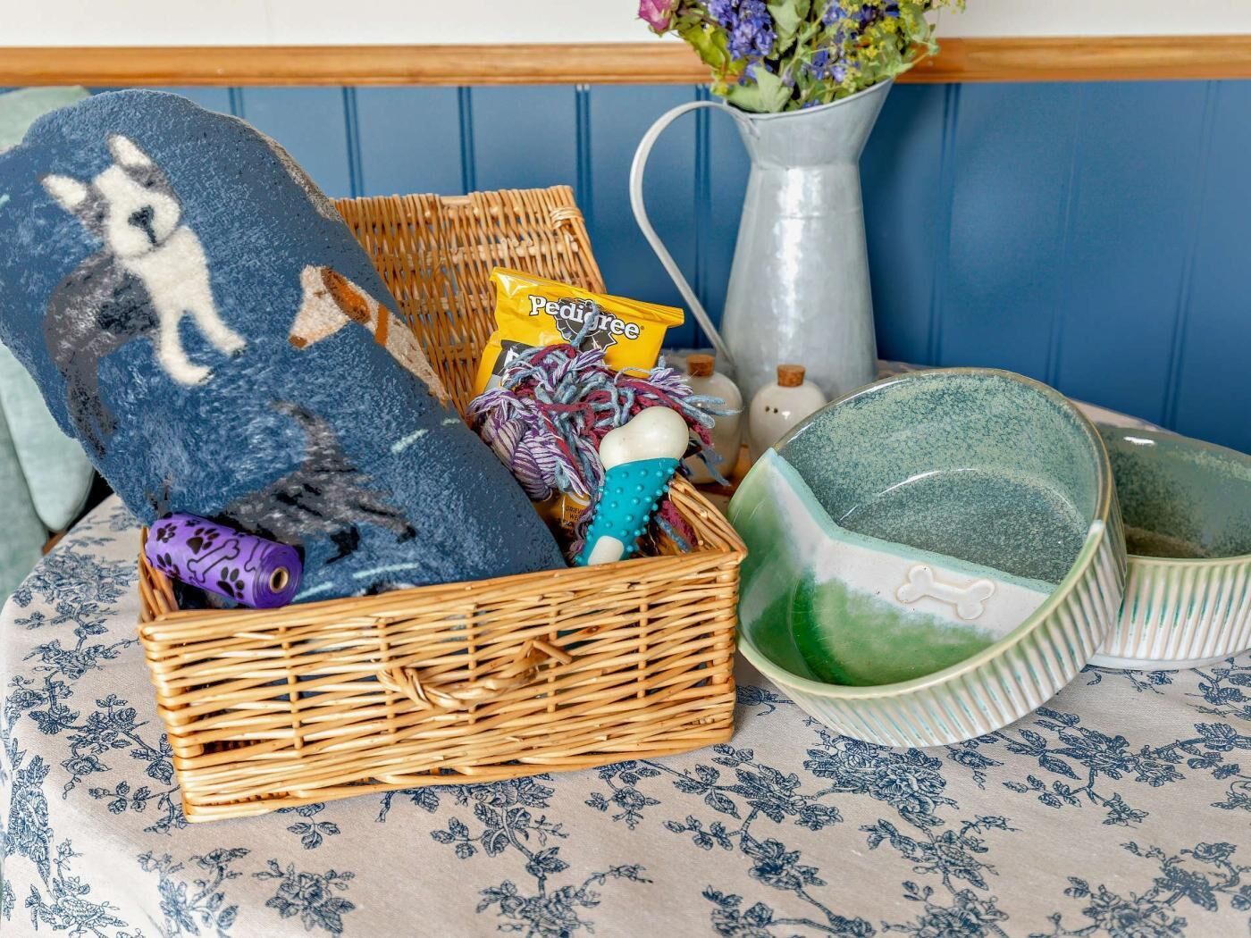 Ein Korbkörbchen mit einem blauen Kissen, eine Vase mit Blumen und eine grüne Keramikschale auf einer gemusterten Tischdecke.