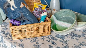 Una cesta de mimbre con un cojín azul, un florero con flores y un cuenco de cerámica verde sobre un mantel estampado.