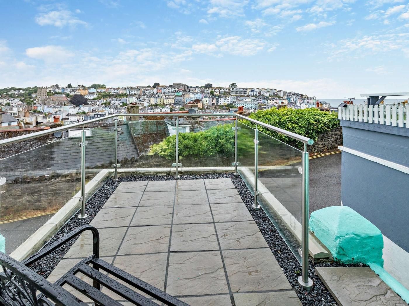 A rooftop terrace with a glass balustrade, a stone floor, and a view of a coastal town.