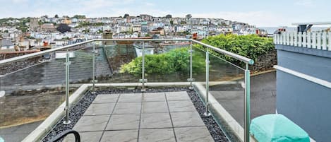 A rooftop terrace with a glass balustrade, a stone floor, and a view of a coastal town.