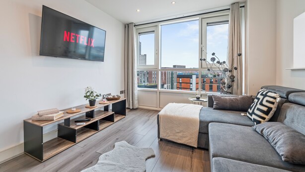 Ein modernes Wohnzimmer mit großem Fernseher, der 'NETFLIX' anzeigt, einem Sofa, einem Couchtisch, einem Bücherregal und einem großen Fenster mit Blick auf die Stadt.