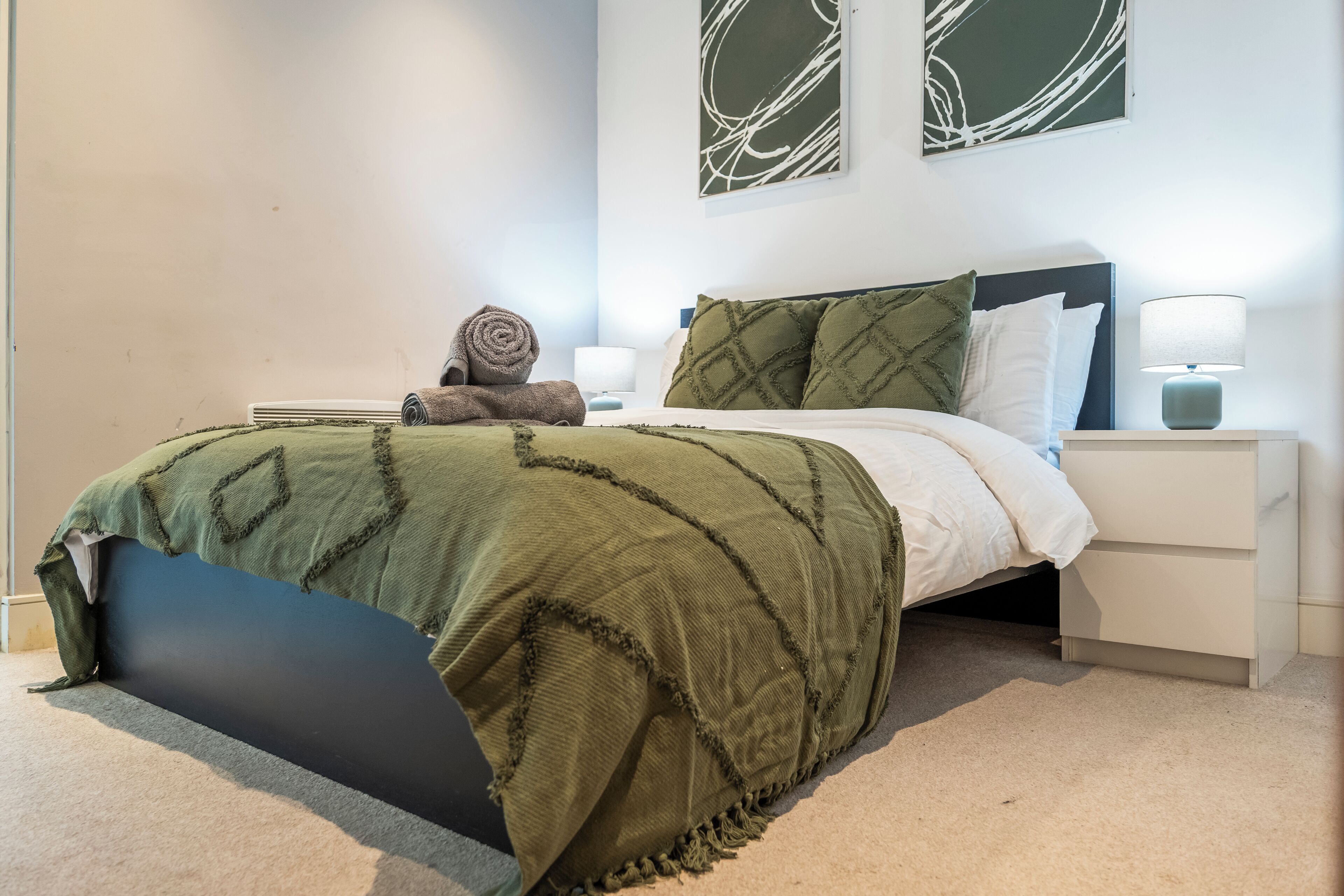 Un lit bien fait, avec une couverture verte, un oreiller et une serviette enroul ée posée sur le côté. Il y a une table de chevet avec une lampe, et une œuvre d’art est accrochée au mur au-dessus du lit.