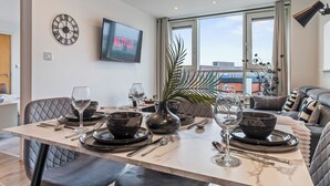 Ein Essbereich mit einem Tisch für vier Personen, einer Uhr, einem Fernseher, der 'NETFLIX' zeigt, und Blick auf die Stadt durch große Fenster.