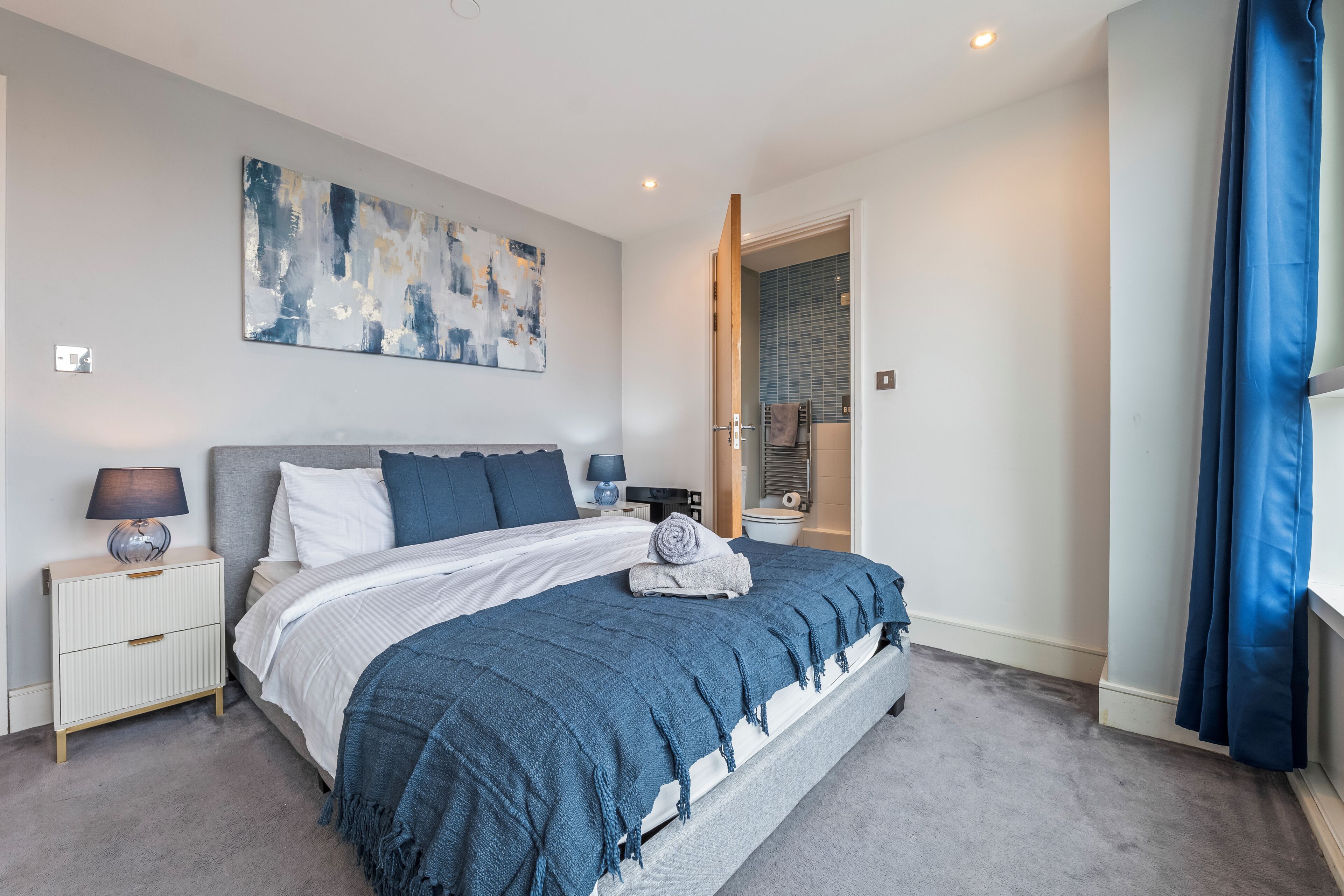 Slaapkamer met een groot bed, nachtkastje, lamp, schilderij aan de muur en een badkamers deur.