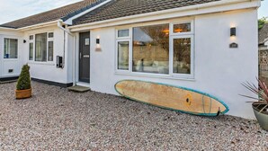 Une maison à deux étages, avec un toit recouvert de tuiles, une planche de surf en bois appuyée contre le mur et une allée gravillonnée.