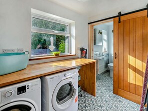 A compact kitchen with a washing machine, a window with a view of greenery, and a sliding door leading to a bathroom. - 1 Bed in Hayle (oc-95418) (Phillack)