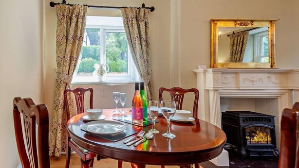 A dining room with a round table, chairs, a fireplace, and a window with curtains. - 1 Bed in Penmaenmawr (oc-95495) (Penmaenmawr)
