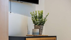 Een flatscreen televisie aan een muur bevestigd, een gevlochten mand met een plant erop een kast, en een houten kast met lades.
